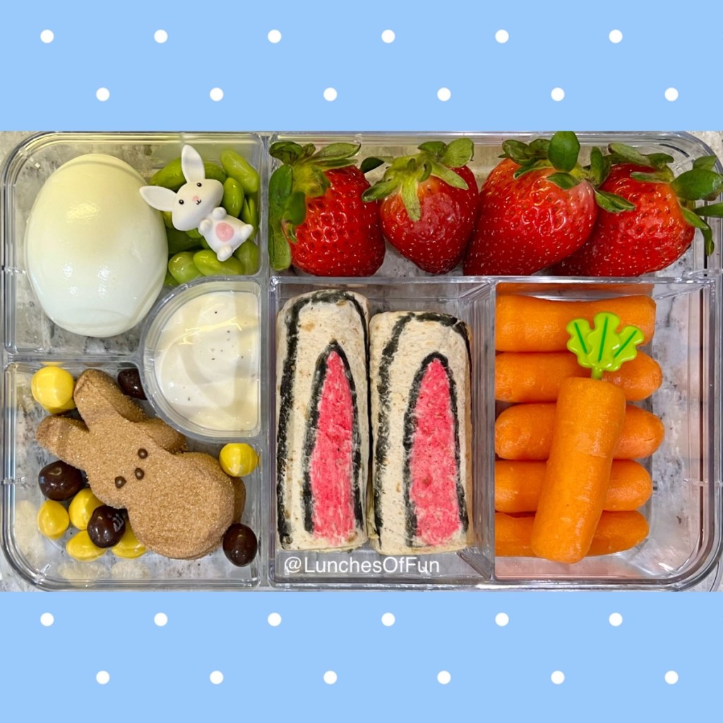 Baby carrot and hummus snack, alongside bunny ears wraps, hard-boiled egg, and chocolate Peep's marshmallow, for a kid's Easter-themed lunchbox