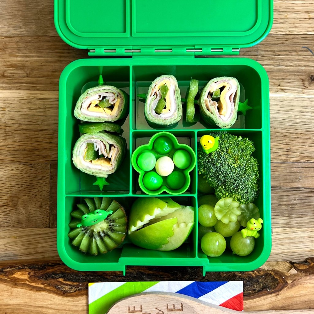 Green lunchbox with delicious spinach wraps rolls, broccoli , kiwi, green graps, and green apple.