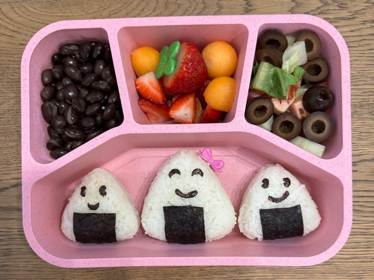 Fun and healthy kids’ lunchbox featuring a family of smiley face onigiri rice balls made with nori in a colorful bento box.