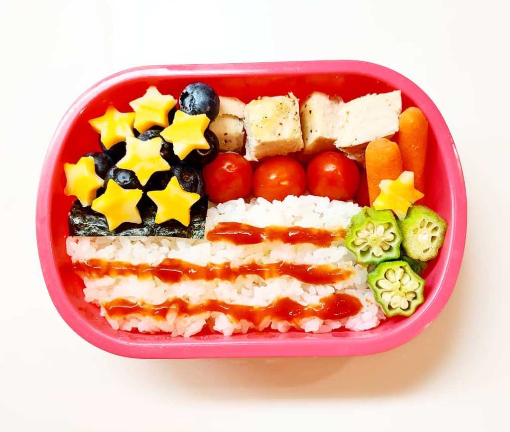 A festive Independence Day lunchbox for kids featuring white rice decorated with ketchup-topped stripes and colorful veggie or cheese accents to mimic the American flag. This easy and patriotic meal is perfect for July 4th celebrations, school lunches, or summer picnics. Find more red, white, and blue lunchbox inspiration at Teuko.com.