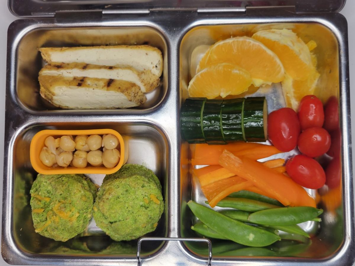 Orange slices, bell pepper strips and other veggies with chicken and mini muffins in a kid's lunchbox
