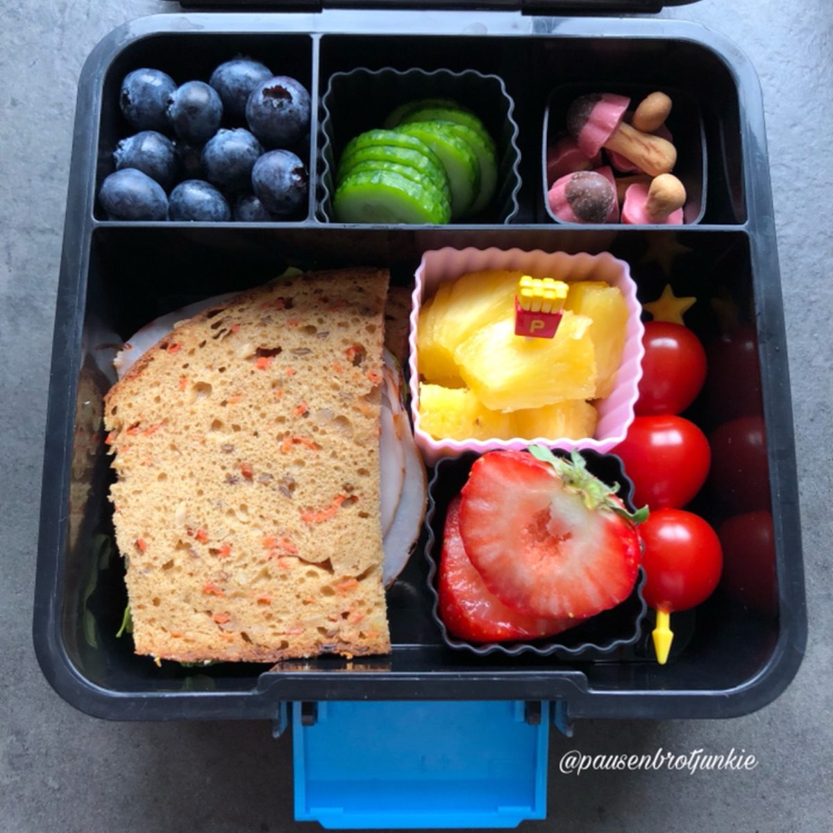 Pineapple chunks, cherry tomatoes, blueberries, cucumber slices, and a carrot cake packed in a lunchbox for a kid's lunch at school