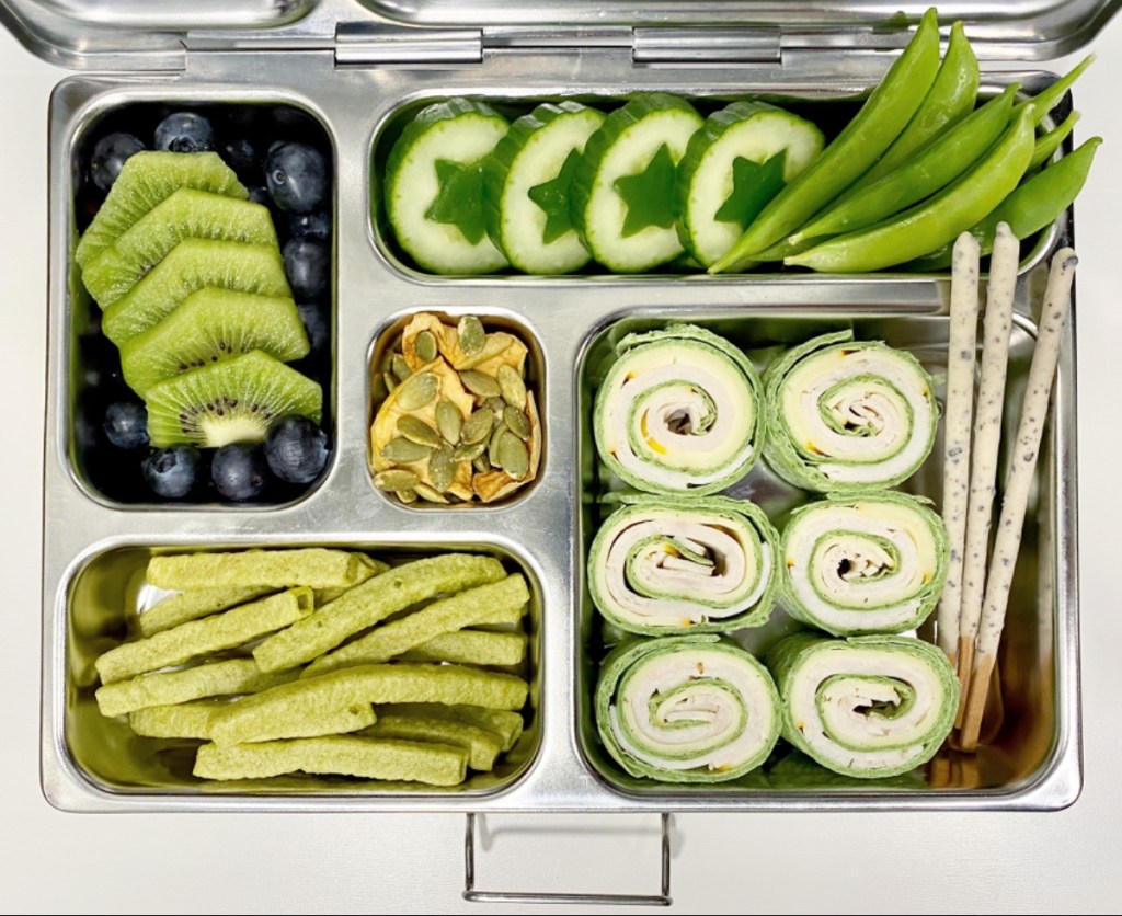 Green color in a kid's lunchbox with kiwi slices, cucumber, snap peas
