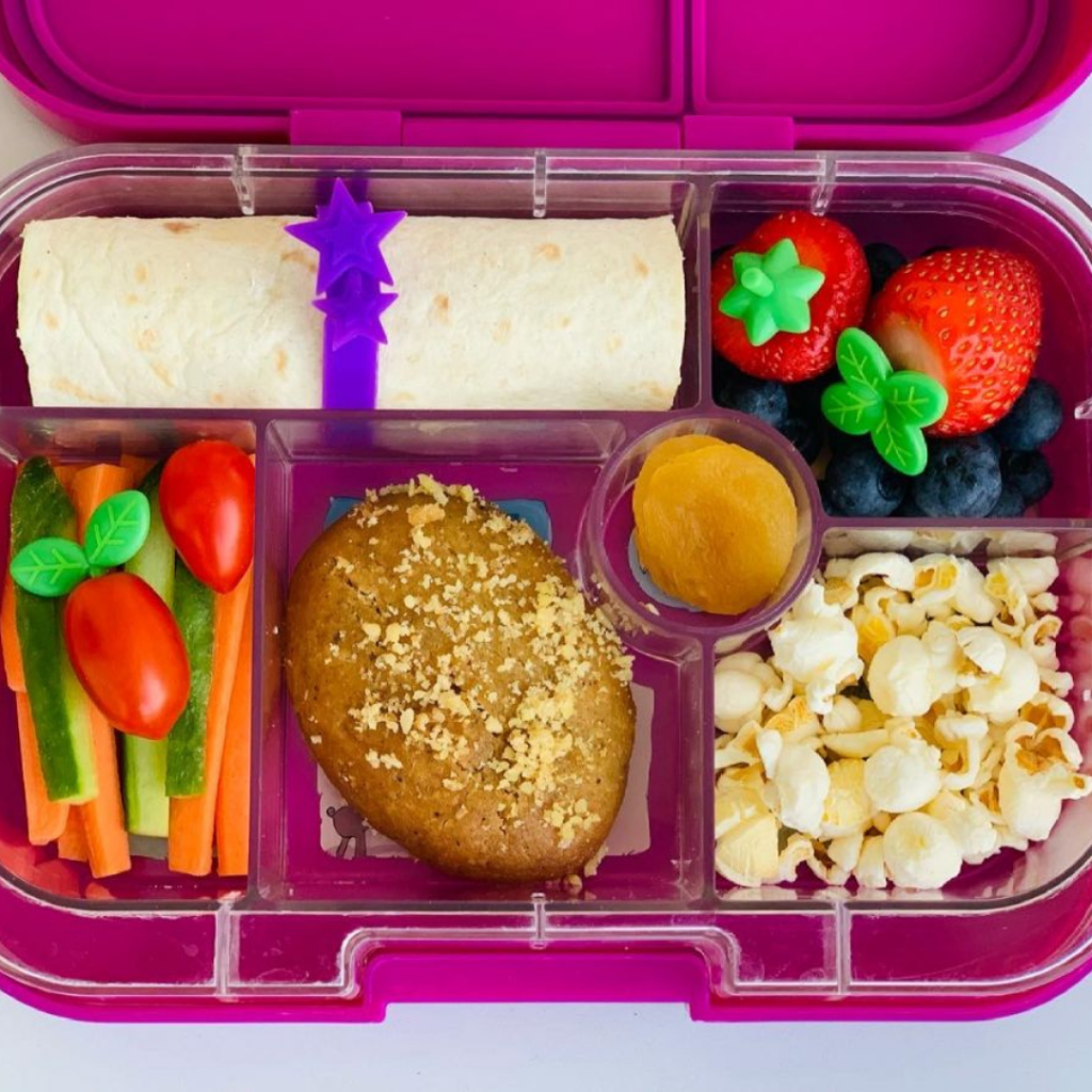 Yumbox with a wrap, carrot and cucumber sticks, banana bread, strawberries and blueberries, dried apricots, and popcorn!