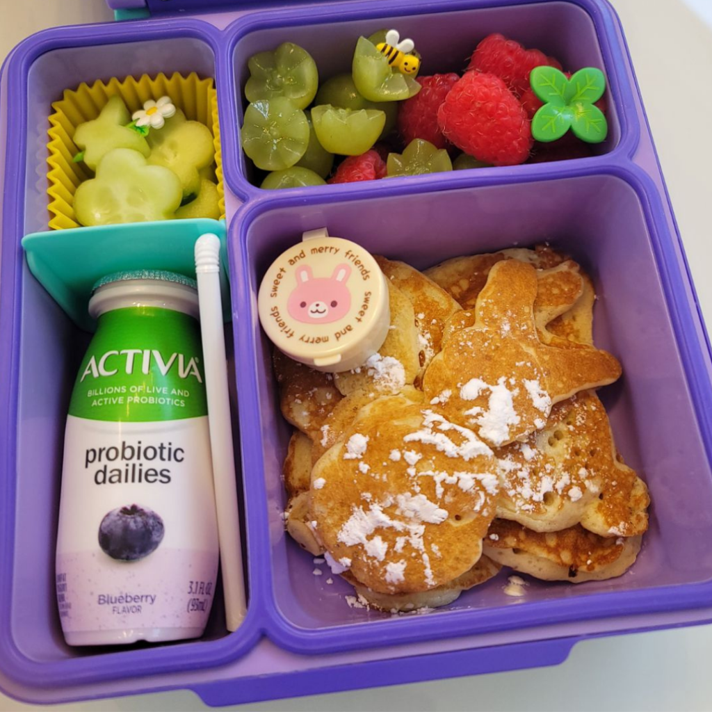 A brunch for her kid with pancakes, eggs, cucumbers, grapes, raspberries, and an Activia probiotic drink.