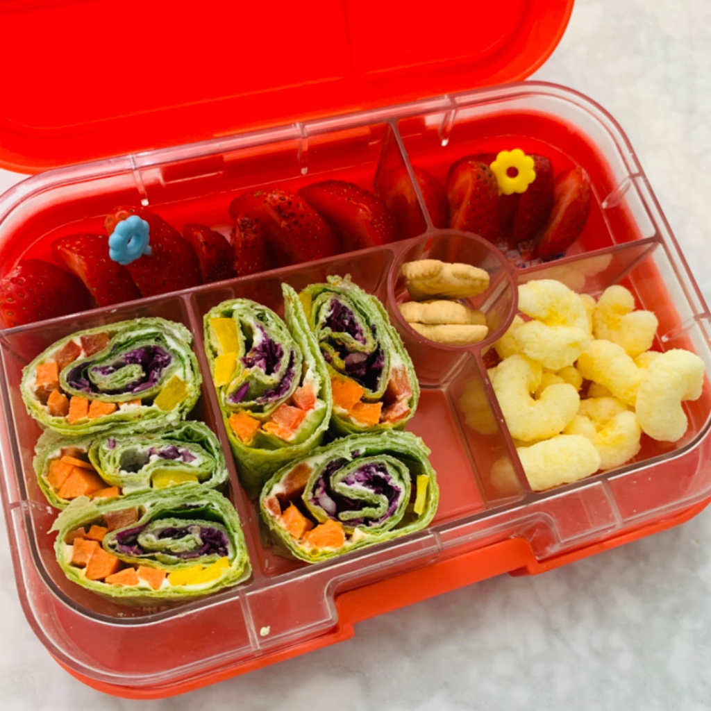 spinach tortilla filled with cream cheese, carrot sticks, bell peppers sliced, and shredded cabbage. You'll note, on the side, cheesy puffs, and strawberries.